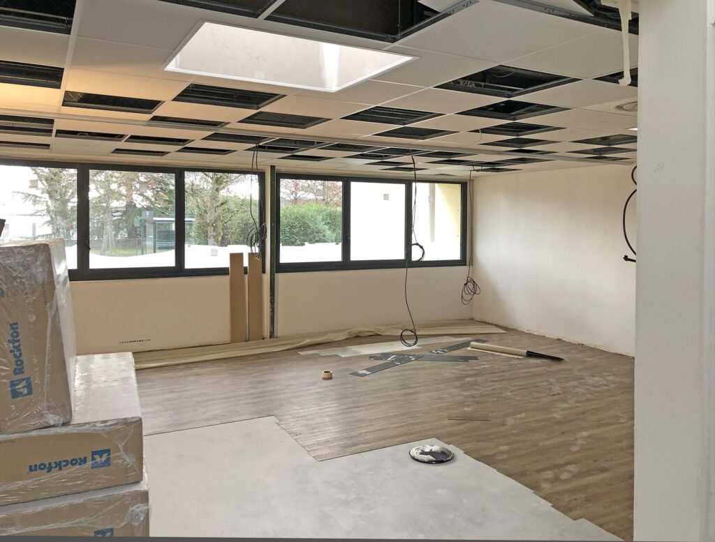 Une pièce non finie avec des panneaux de plafond apparents, des murs nus, de grandes fenêtres et des boîtes d'isolation empilées sur la gauche. L'installation du revêtement de sol semble être en cours.