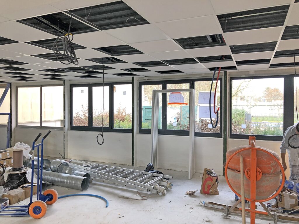 Espace de bureau en cours de construction avec câblage apparent, échelles, conduits de ventilation, outils et un plafond suspendu partiellement installé. De grandes fenêtres apportent de la lumière naturelle.