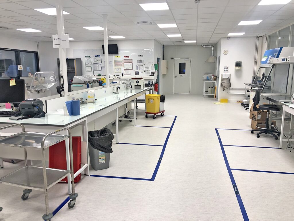 Un laboratoire propre et moderne avec des établis, du matériel et des poubelles. Des rubans bleus délimitent des zones sur le sol. Le laboratoire est bien éclairé et organisé, les panneaux et les postes de sécurité sont visibles.
