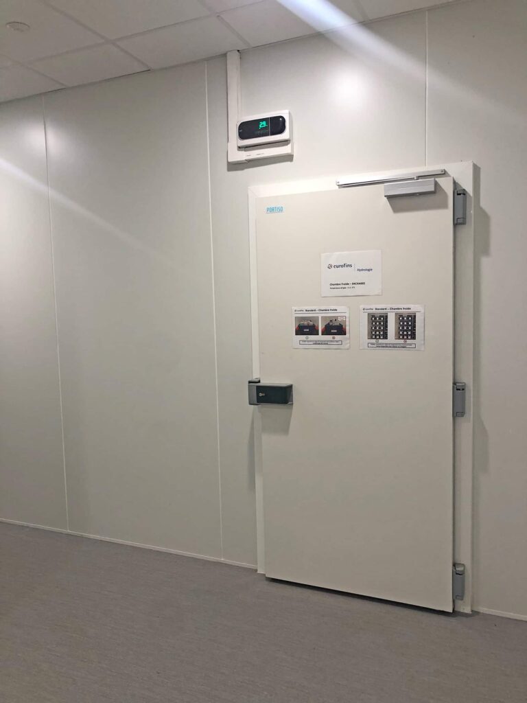 Une porte de sécurité blanche fermée avec des instructions et un clavier, placée dans un mur blanc simple sous un petit écran numérique.