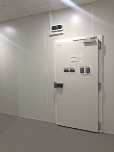 Une porte blanche, d'aspect industriel, dotée d'un clavier et de panneaux d'instructions, est encastrée dans un mur blanc ordinaire, sous un affichage numérique de la température.