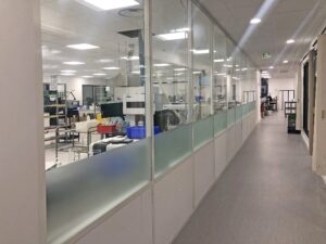 Un couloir aux parois vitrées révèle un espace de travail de laboratoire moderne rempli d'équipements, de tables et de bacs de rangement, avec un éclairage fluorescent au-dessus de la tête.