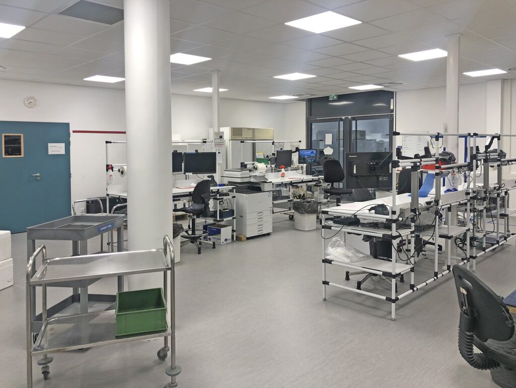 Un espace de travail de laboratoire propre et moderne avec des bureaux, des ordinateurs, du matériel médical, des chariots et des chaises disposés dans un environnement bien éclairé et organisé.