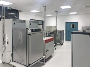 Une salle de laboratoire avec plusieurs grands équipements en acier inoxydable sur roulettes, un éclairage zénithal, une porte bleue et un intérieur blanc et propre.