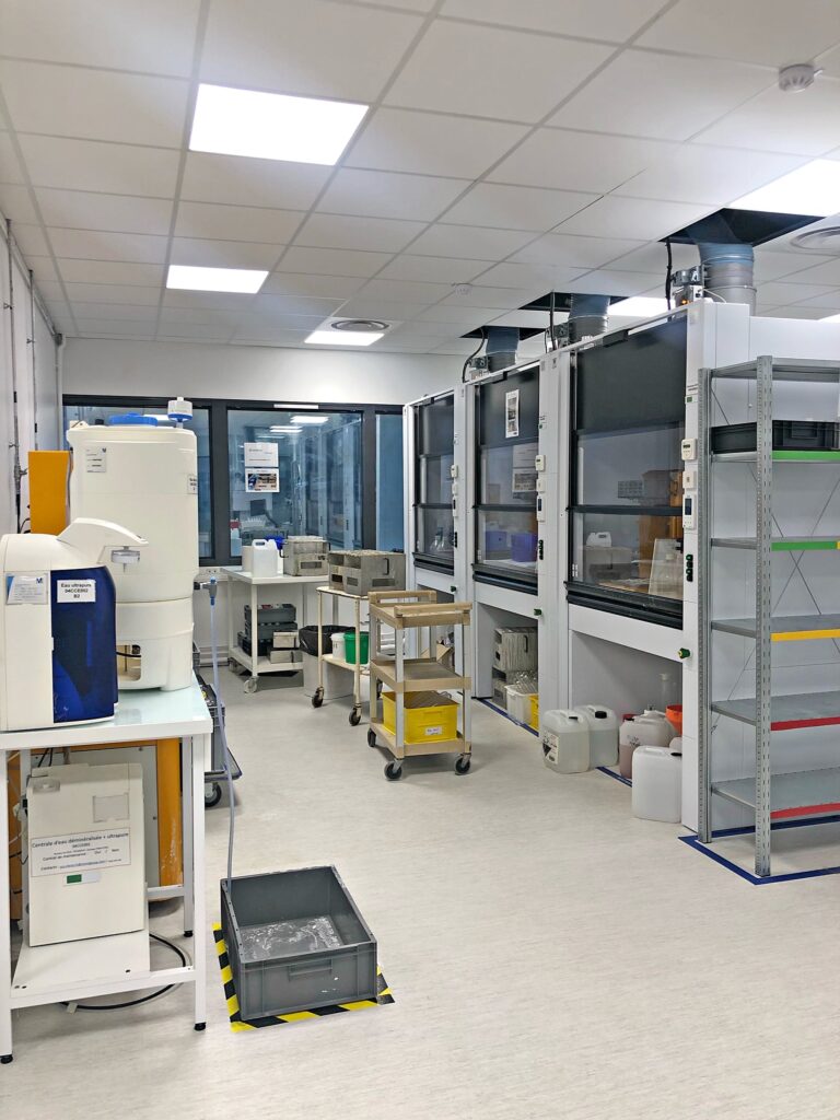 Un laboratoire moderne avec des postes de travail, des équipements, des étagères de rangement, des chariots et des armoires de sécurité sous un éclairage zénithal intense.
