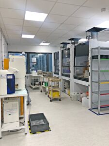 Un laboratoire propre et organisé avec des postes de travail, des étagères, du matériel et des conteneurs de stockage de sécurité sous un éclairage zénithal lumineux.
