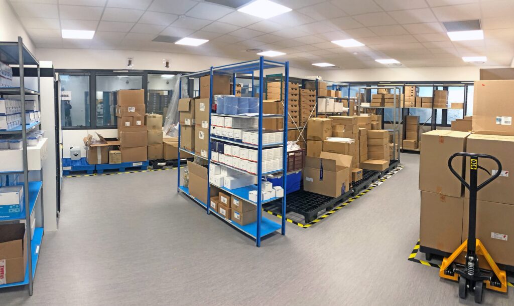 Intérieur d'un entrepôt avec des étagères et des palettes empilées avec des cartons, des bacs de stockage et un transpalette au sol. L'éclairage est clair et l'agencement organisé.