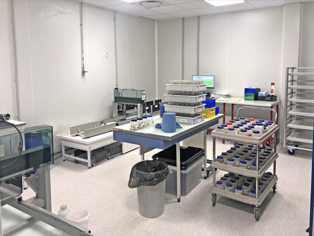 Une salle de laboratoire propre avec des établis, des plateaux de bouteilles de laboratoire, des étagères, une poubelle et du matériel scientifique bien rangé.