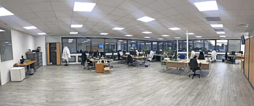 Bureau moderne et spacieux avec des bureaux, des chaises, des ordinateurs, des classeurs et de grandes fenêtres ; des plafonniers fluorescents éclairent la pièce.