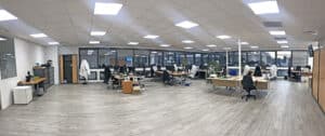 Bureau spacieux, moderne et ouvert, avec des bureaux, des chaises, des ordinateurs, de grandes fenêtres et un éclairage zénithal puissant ; personne n'est présent.