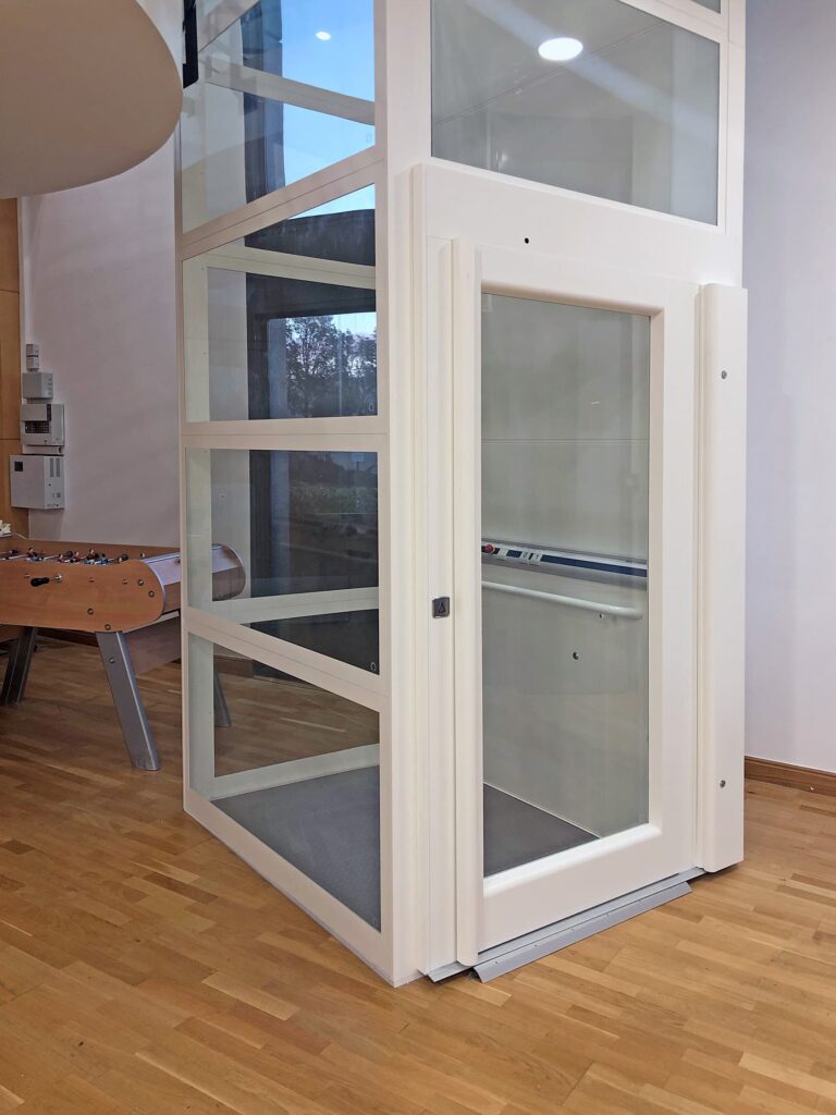 Un ascenseur domestique moderne en verre avec un cadre blanc est installé à côté d'une table de baby-foot sur un sol en bois dans une pièce bien éclairée.