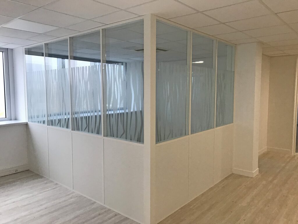 Une cloison de bureau moderne avec des panneaux en verre dépoli et un encadrement blanc, séparant une partie de la pièce près de grandes fenêtres et d'un sol de couleur claire.