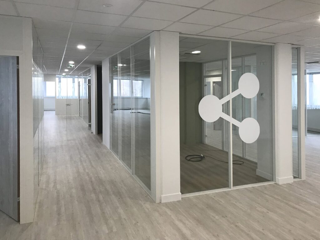 Couloir de bureau moderne avec des murs en verre, un sol en bois clair et une grande icône de réseau blanche sur la fenêtre d'une salle de réunion.