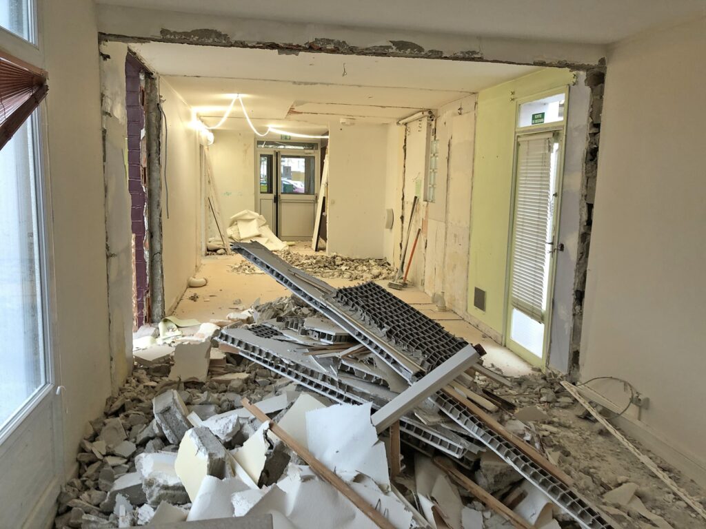 Une pièce en cours de rénovation avec des débris, des cloisons sèches cassées et des montants métalliques éparpillés sur le sol ; les murs sont partiellement démolis.