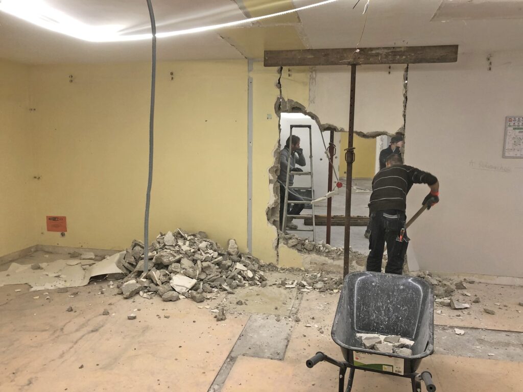 Un ouvrier utilise un outil pour démolir un mur, créant un grand trou ; des débris et une brouette se trouvent sur le sol.