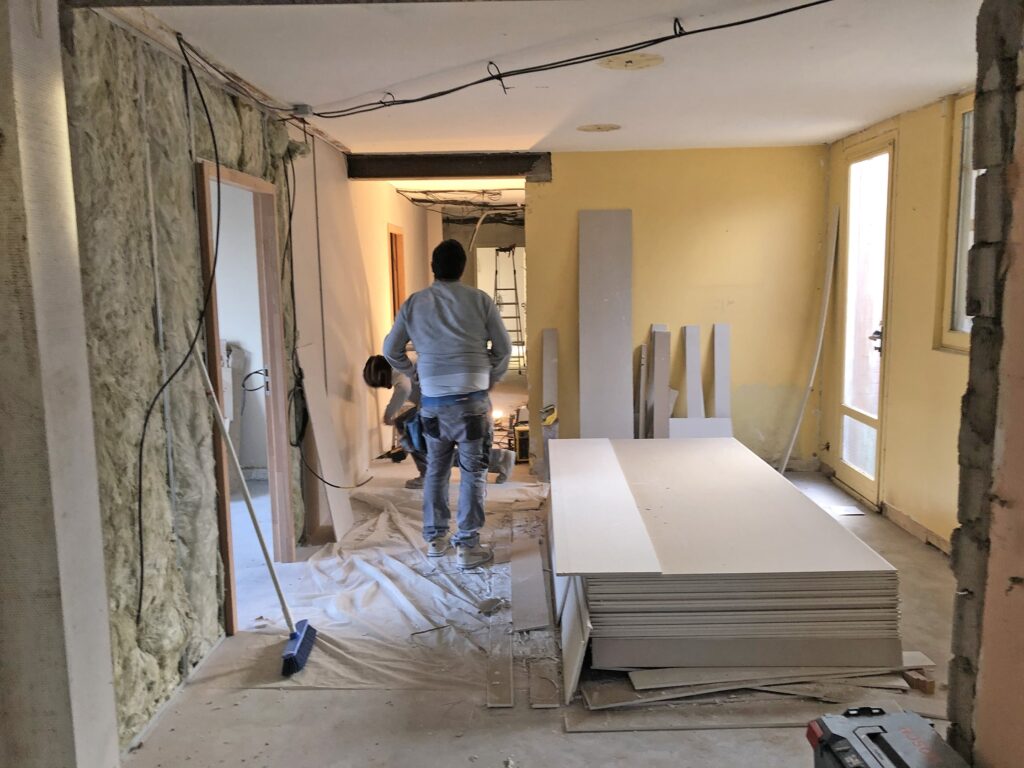 Deux personnes travaillant dans une pièce en cours de rénovation, avec de l'isolant sur un mur, des plaques de placoplâtre empilées sur le sol et des outils et matériaux de construction éparpillés.