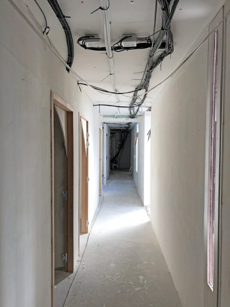 Couloir non aménagé avec des câbles apparents au plafond, des murs non peints et des portes menant à des pièces en construction.