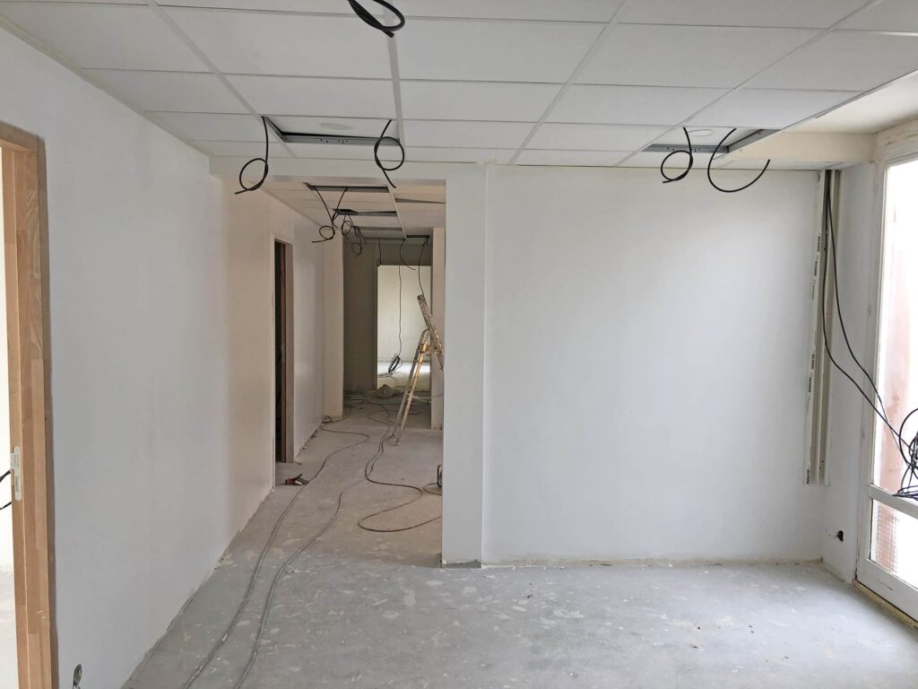 Une pièce en cours de rénovation avec des murs blancs, des sols non finis, des fils électriques apparents qui pendent du plafond et des matériaux de construction visibles à l'arrière-plan.