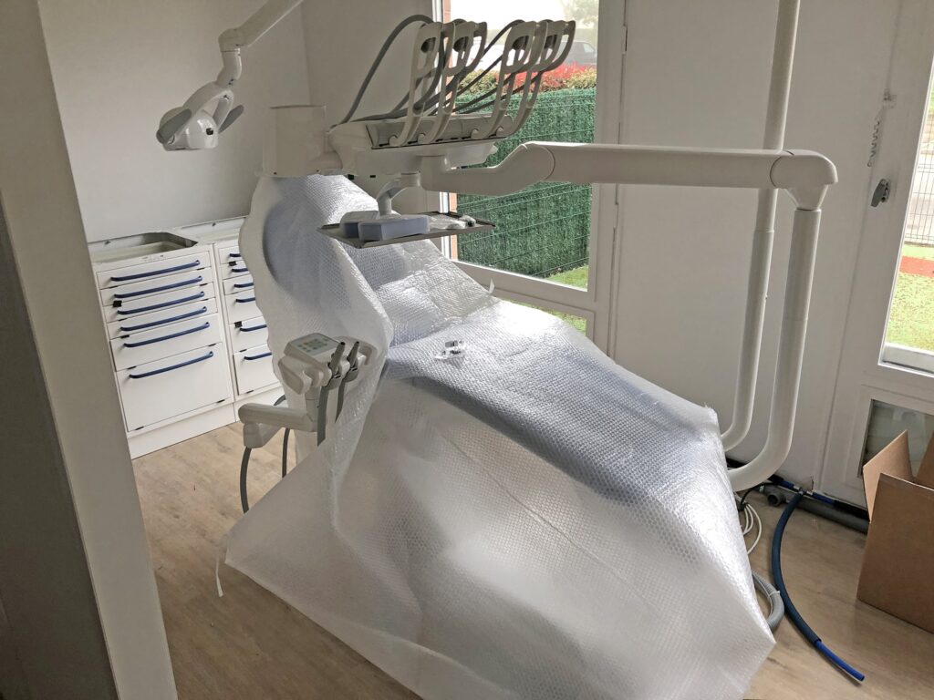 Un fauteuil de dentiste recouvert de papier bulle est placé dans un cabinet dentaire clair et propre, doté d'armoires et d'une grande fenêtre.