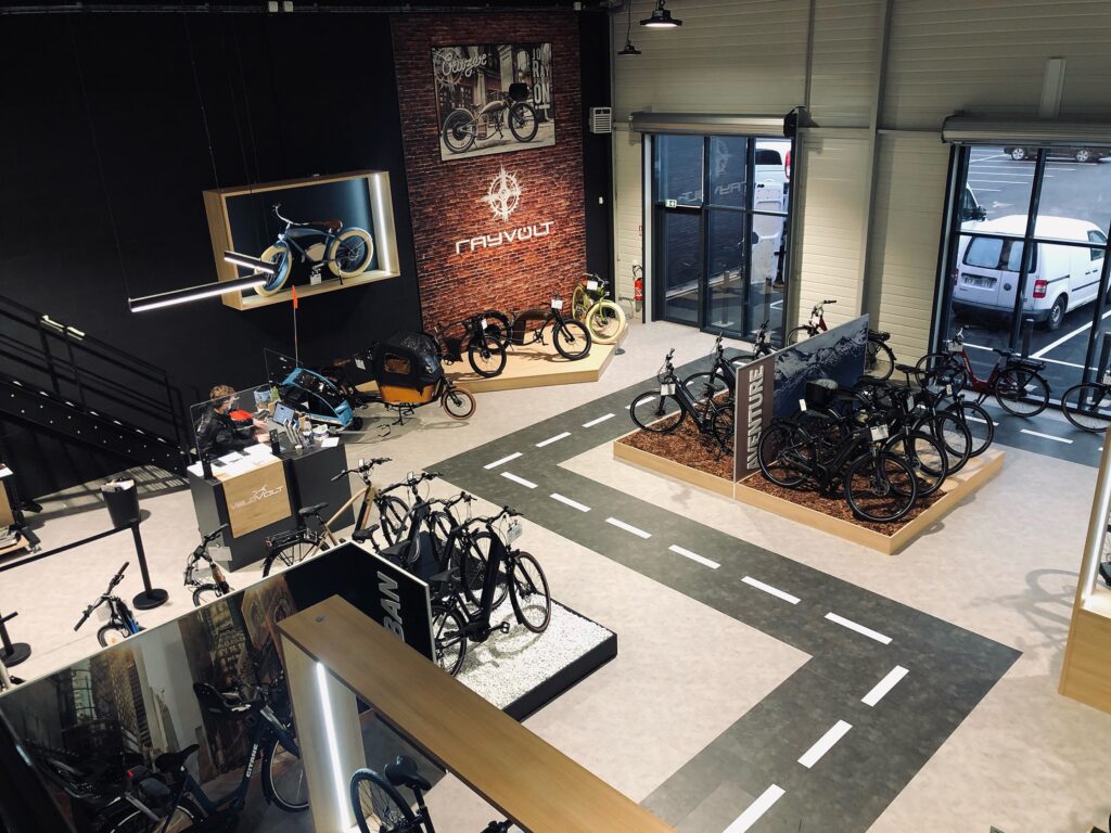Salle d'exposition de bicyclettes avec différents vélos exposés, une route fictive peinte sur le sol et de grandes fenêtres laissant entrer la lumière naturelle.