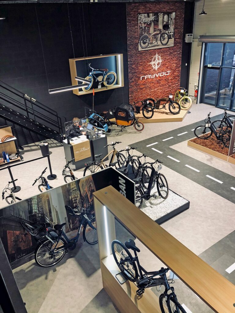 Intérieur d'un magasin de vélos moderne avec plusieurs vélos en exposition, une route fictive pour les essais et de grandes affiches sur les murs. Une personne travaille au comptoir.