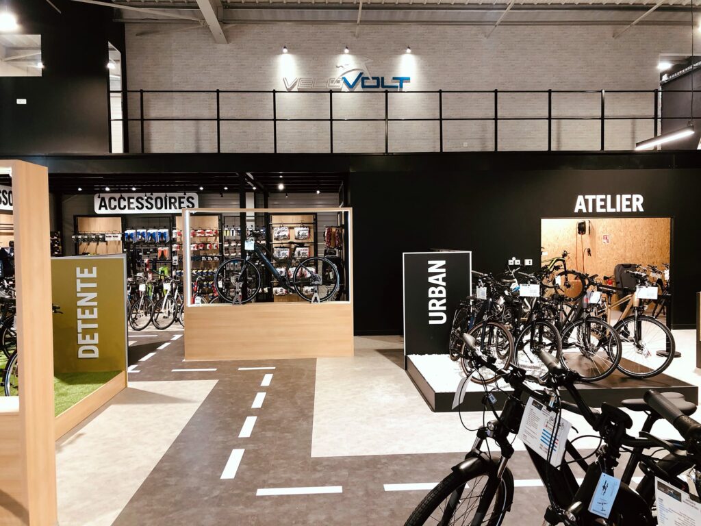 Intérieur d'un magasin de vélos moderne avec des sections étiquetées pour les accessoires, les vélos urbains, l'atelier et l'espace détente ; divers vélos et équipements de cyclisme sont exposés.