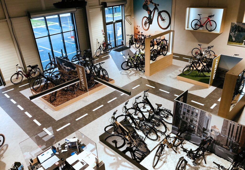 Vue large de l'intérieur d'un magasin de vélos moderne avec divers vélos en exposition, un décor sur le thème du cyclisme et un sentier balisé ressemblant à une route sur le sol.