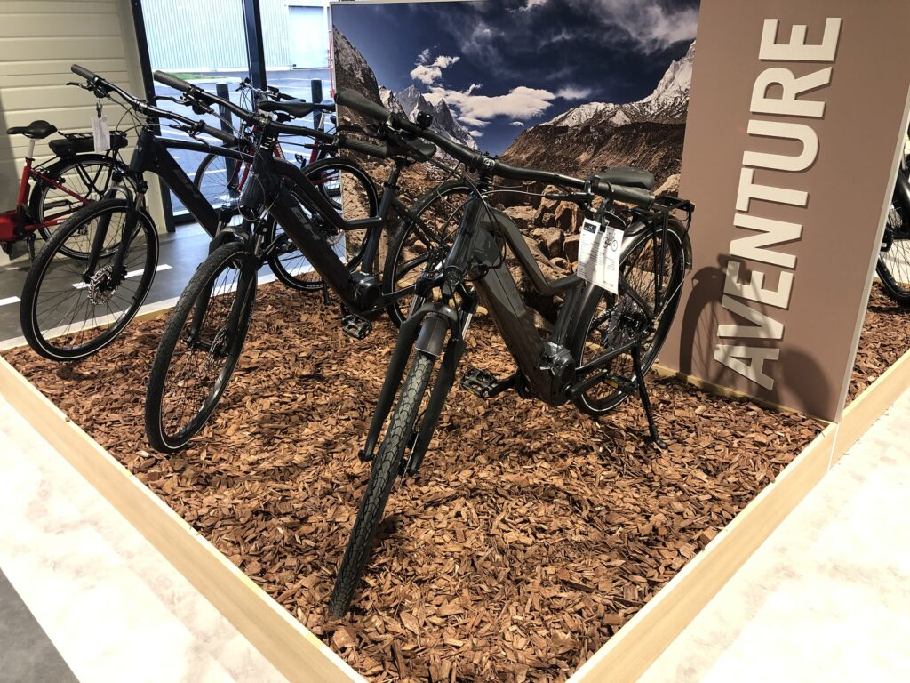 Une exposition de vélos électriques sur des copeaux de bois devant un paysage de montagne, avec un grand panneau indiquant "AVENTURE".