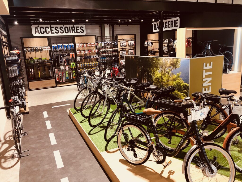 Magasin de vélos intérieur avec des rangées de vélos en exposition, des accessoires sur des étagères murales et des panneaux indiquant "Accessoires" et "Bargerie" au-dessus des étagères.