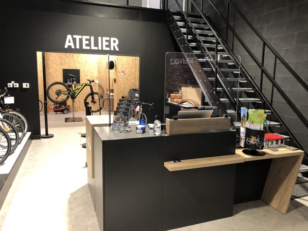 Intérieur d'un magasin de bicyclettes moderne avec une réception, un escalier et un atelier portant la mention "Atelier" à l'arrière-plan. Des bicyclettes et des outils de réparation sont visibles.