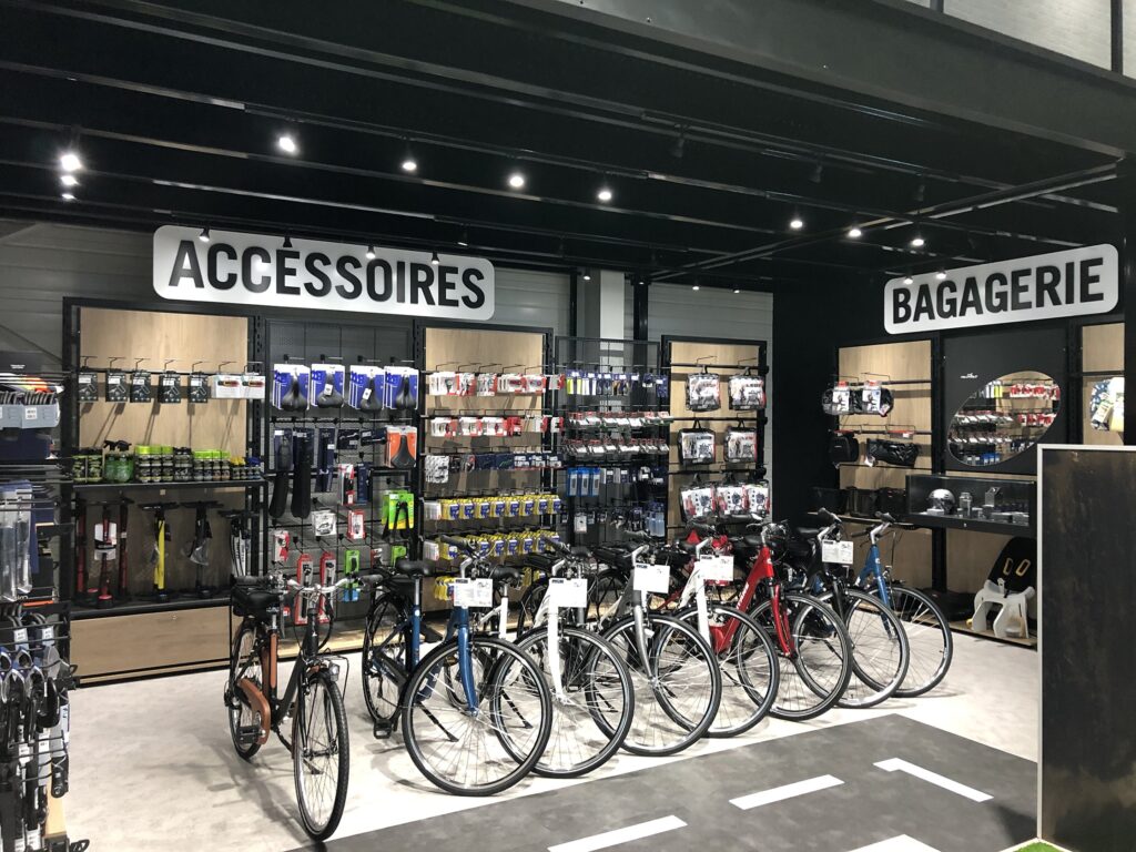 Un magasin de vélos avec plusieurs vélos devant et des étagères d'accessoires et de sacs étiquetés "Accessoires" et "Bagagerie" à l'arrière-plan.