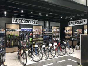 Un magasin de vélos avec plusieurs vélos devant et des étagères d'accessoires et de sacs étiquetés "Accessoires" et "Bagagerie" à l'arrière-plan.