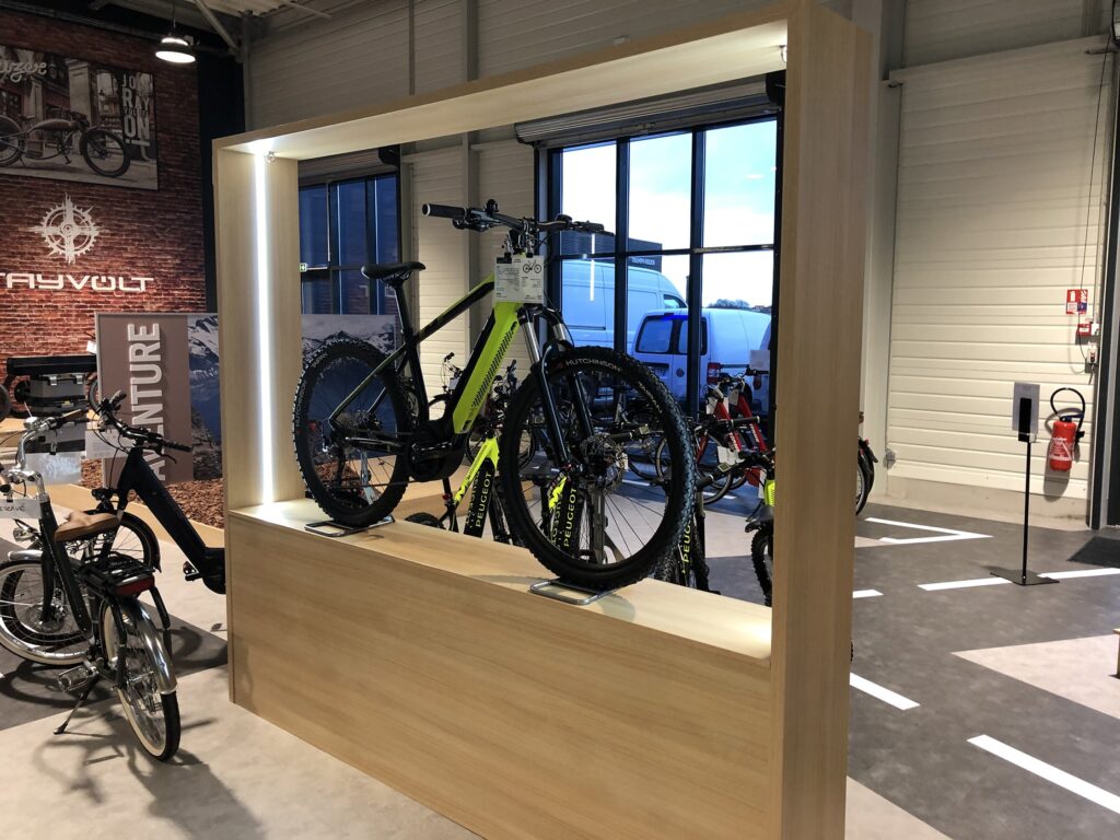 Un vélo tout-terrain vert présenté sur une plate-forme en bois dans une salle d'exposition, avec d'autres vélos visibles à l'arrière-plan.