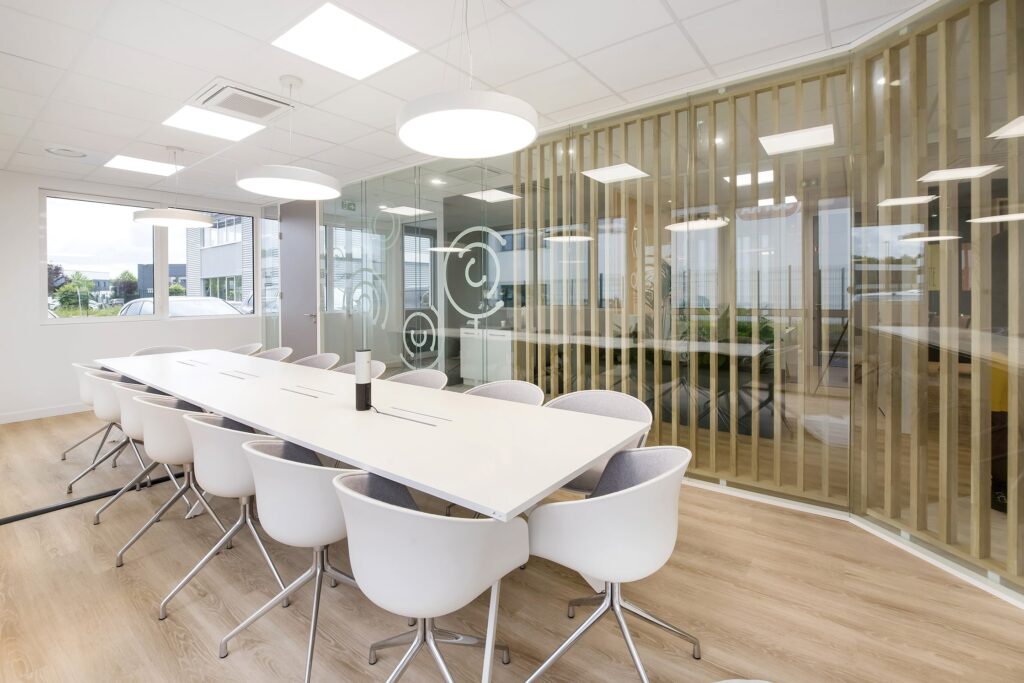 Salle de conférence moderne avec une longue table blanche, des chaises blanches, des lampes suspendues, des murs en verre et la lumière naturelle provenant de grandes fenêtres.