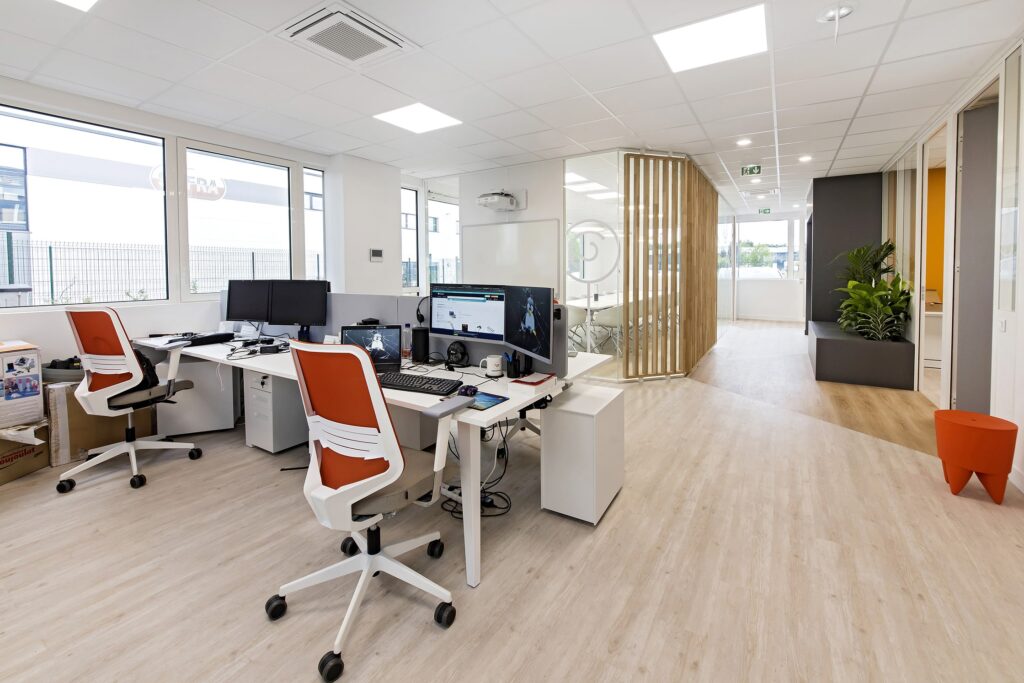 Espace de bureau moderne avec bureaux, ordinateurs et chaises orange ; murs blancs, grandes fenêtres, parquet en bois clair et plantes en pot.