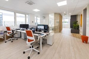 Espace de bureau moderne avec bureaux, ordinateurs et chaises orange ; murs blancs, grandes fenêtres, parquet en bois clair et plantes en pot.