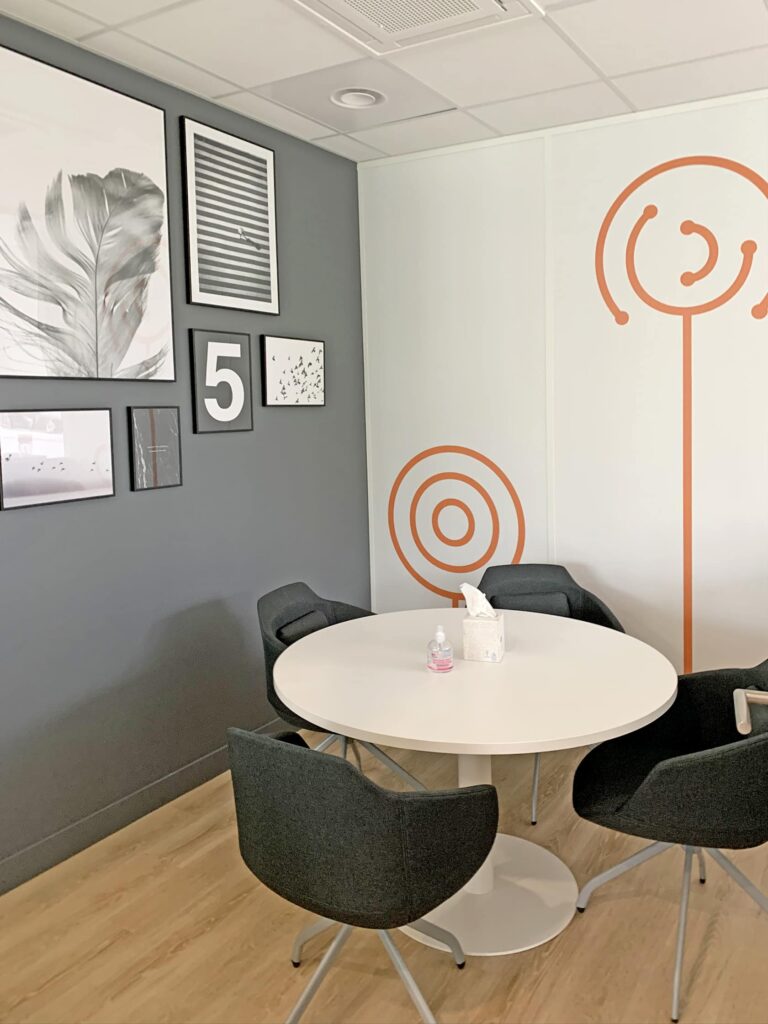 Un petit espace de réunion avec une table ronde blanche, quatre chaises noires, de l'art mural, des reproductions encadrées et des motifs abstraits orange sur des murs blancs.