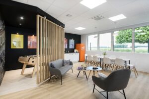 Salle de pause moderne avec chaises longues, baby-foot, table à manger, kitchenette, réfrigérateur orange, œuvres d'art murales et grandes fenêtres donnant sur la verdure.