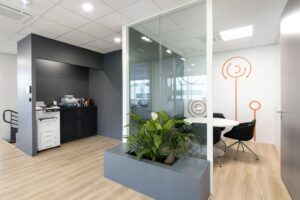Espace de bureau moderne avec une petite salle de réunion fermée par une vitre, une table ronde blanche avec des chaises noires, des plantes d'intérieur et des équipements de bureau en arrière-plan.