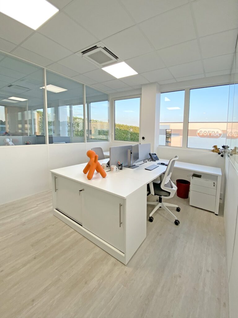 Bureau moderne avec des meubles blancs, deux écrans d'ordinateur sur un bureau, un ornement en forme de chien orange et de grandes fenêtres laissant entrer la lumière naturelle.