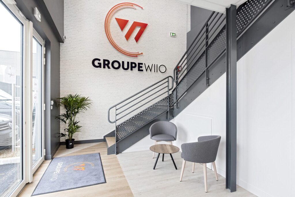 Hall d'entrée d'un bureau moderne avec deux chaises grises, une petite table ronde, une plante en pot, un escalier et le logo "GROUPE WIO" sur un mur de briques blanches.