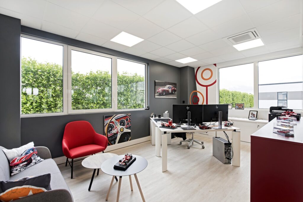 Bureau moderne avec deux bureaux, des ordinateurs, une chaise rouge, un canapé gris, des tableaux muraux représentant des voitures et de grandes fenêtres laissant entrer la lumière naturelle.