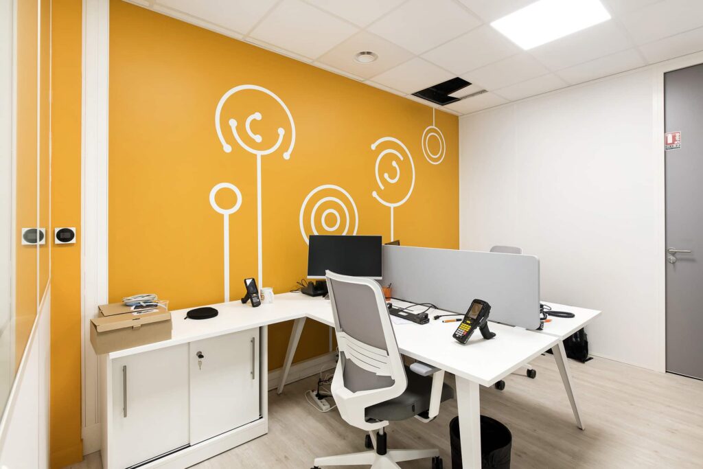 Bureau moderne avec deux bureaux blancs, des chaises, des ordinateurs et un mur d'accent jaune avec des motifs abstraits blancs ; dalle de plafond partiellement ouverte.