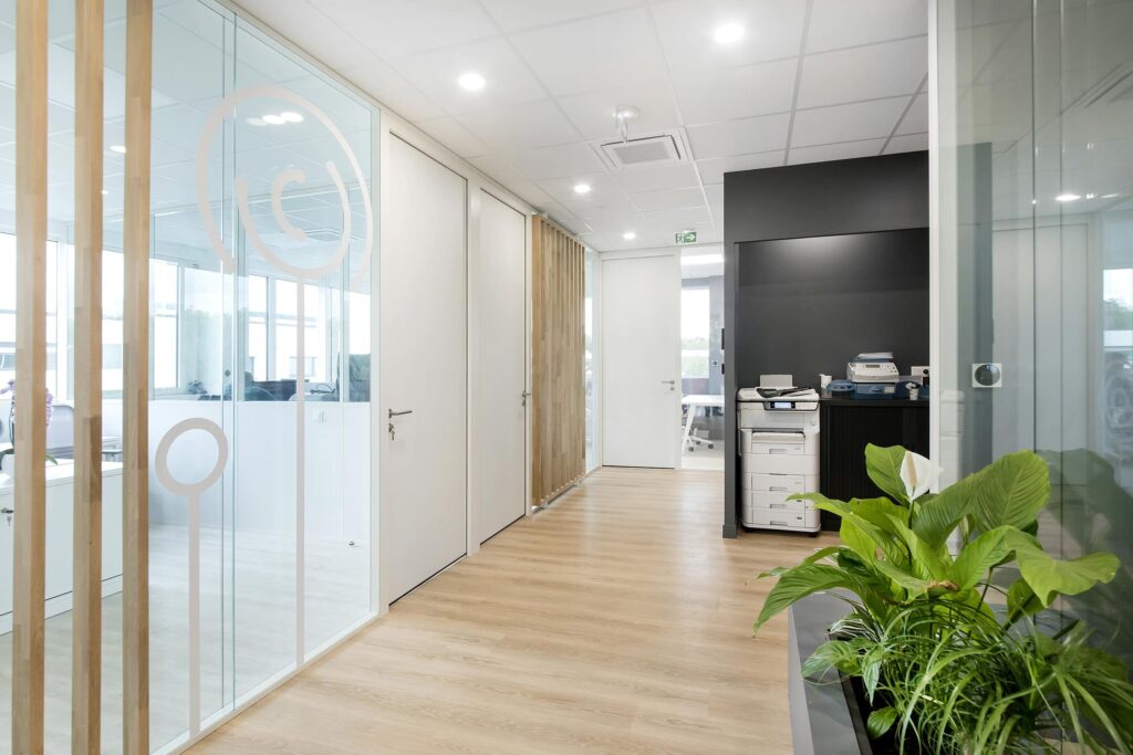 Couloir de bureau moderne avec des murs en verre, un sol en bois, des portes blanches, une station d'impression, des plantes d'intérieur et des plafonniers lumineux.