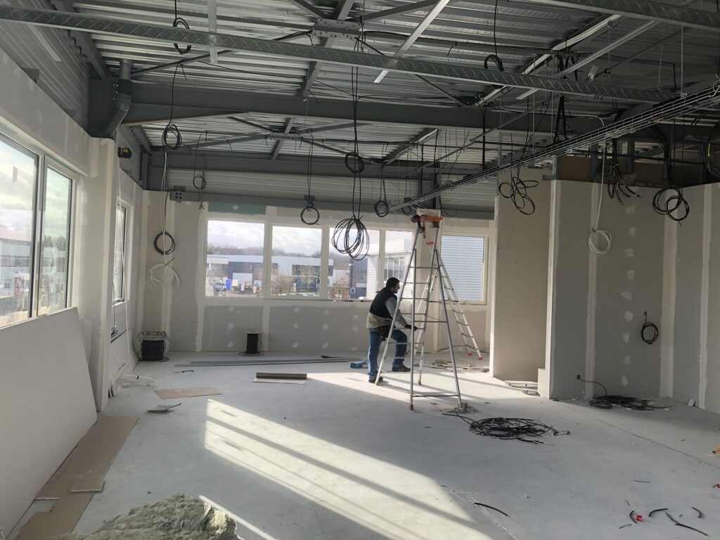 Une personne se tient sur une échelle et travaille à l'intérieur d'une pièce en construction, avec des câbles exposés, des murs inachevés et des matériaux de construction éparpillés sur le sol.