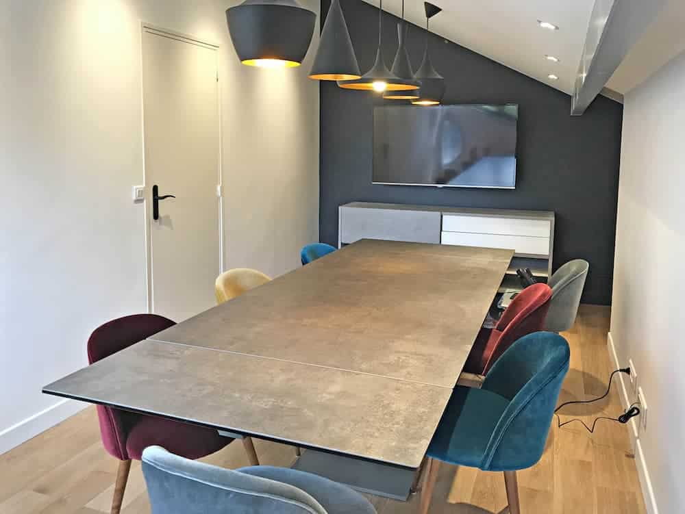 Salle de conférence moderne avec une longue table rectangulaire, des chaises en velours coloré, des lampes suspendues, une télévision murale et un buffet contre un mur d'accent sombre.