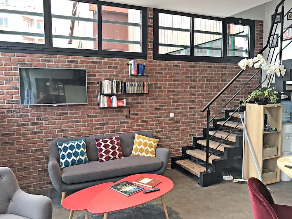 Salon moderne avec un mur de briques apparentes, un canapé gris avec des coussins colorés, une télévision murale, une bibliothèque, un escalier et une table basse rouge contenant des livres.