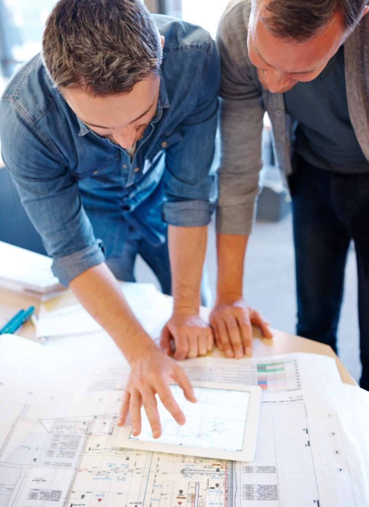 Deux hommes debout examinent des plans d'architecture sur un bureau, l'un d'eux montrant une tablette affichant des plans.