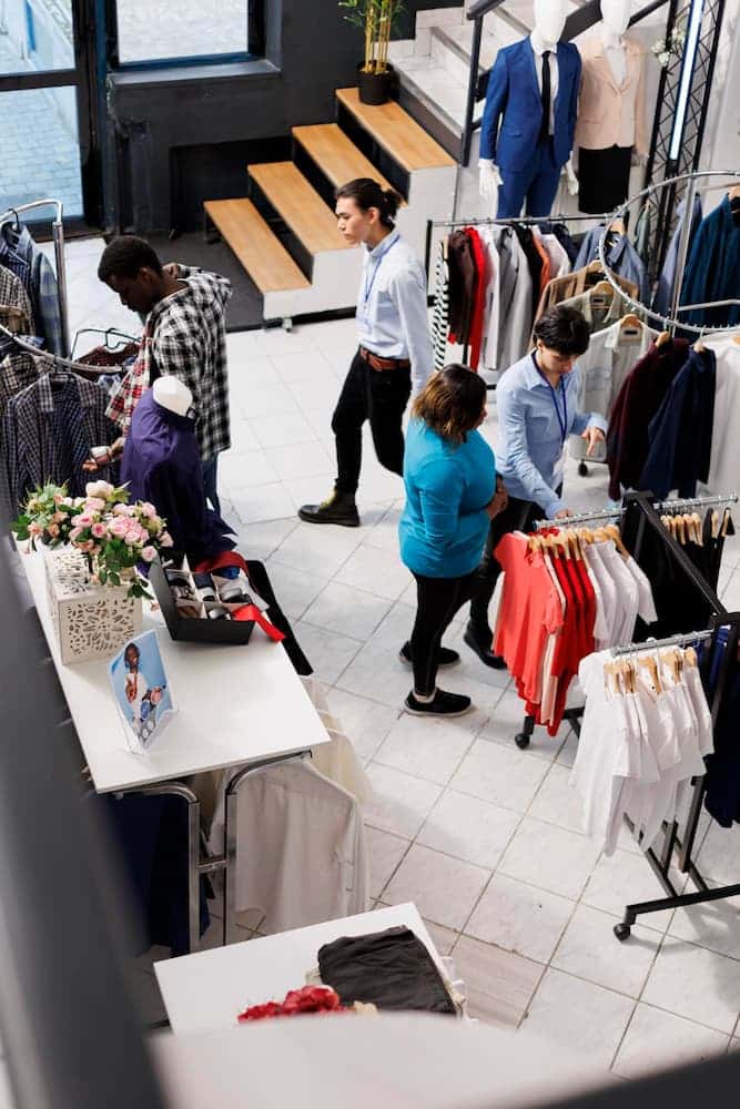 Des personnes font du shopping et interagissent avec le personnel à l'intérieur d'un magasin de vêtements, avec des étagères de vêtements et des mannequins en exposition.