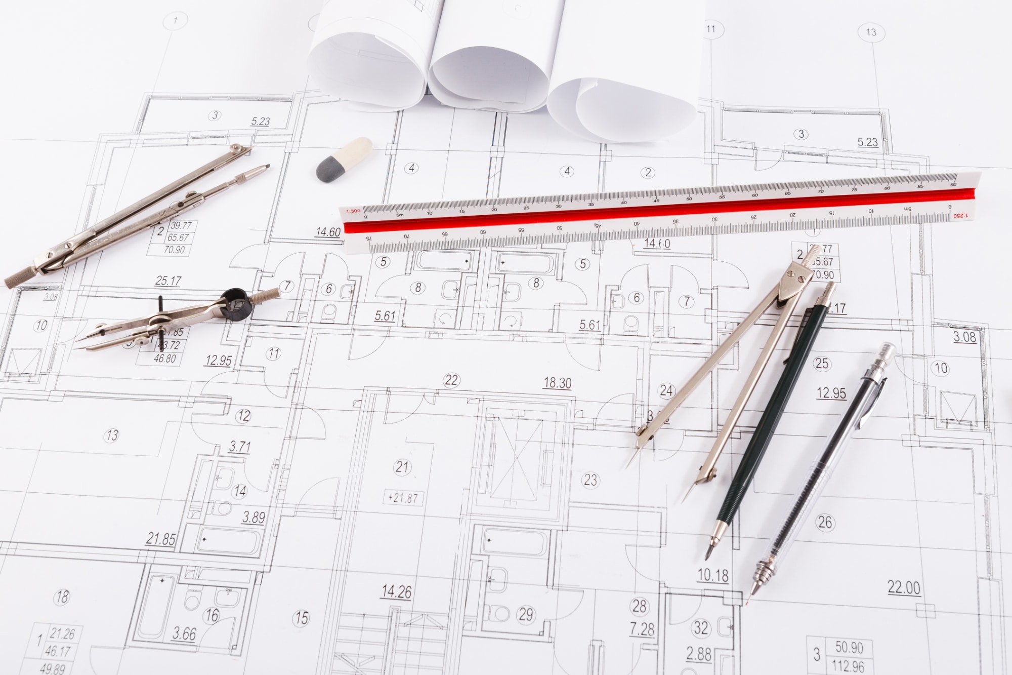 Plan d'architecture posé sur un bureau d'aménagement de l'espace, avec des outils de dessin, notamment deux compas, une règle graduée, une gomme, un crayon et trois plans techniques enroulés.